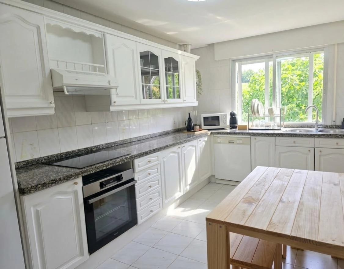 Vista de la cocina reformada de Casa Buen Camino en Arzúa. Se observan muebles y armarios superiores de color blanco impecable, encimera de granito veteado, placa de inducción, horno y microondas encastrados. Una mesa de madera clara con bancos ocupa el primer plano y una ventana al fondo ilumina toda la estancia con luz natural y vistas al jardín.