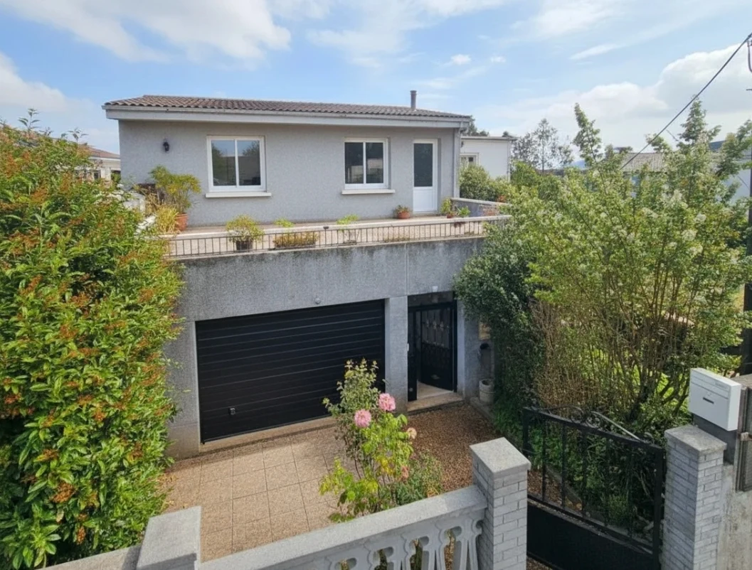 Fachada exterior de Casa Buen Camino en Arzúa. Se observa la casa rural de dos plantas con pared gris granítica, un gran garaje de persiana negra en la planta baja, una entrada peatonal con reja negra y una amplia terraza soleada en la planta alta decorada con plantas en macetas. Un jardín cuidado con arbustos verdes y un rosal rosa enmarca el acceso peatonal de grava.