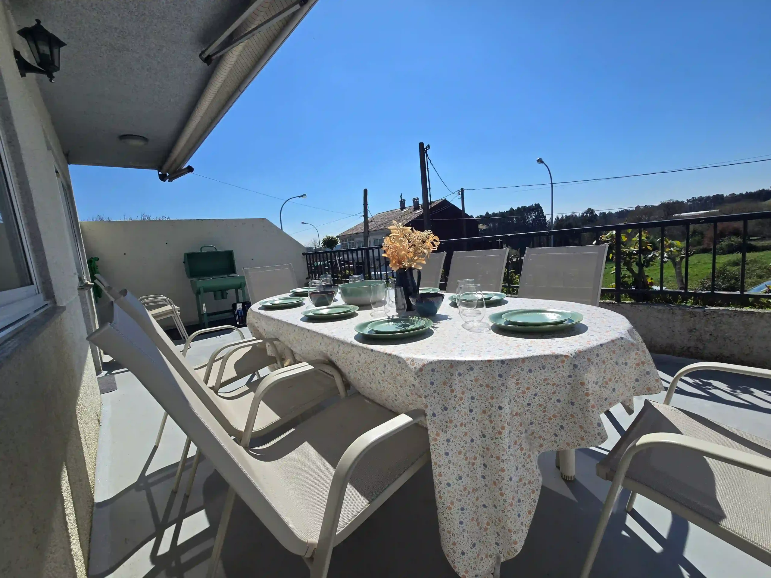 Amplia terraza exterior en Casa Buen Camino, Arzúa, equipada con una mesa grande de comedor para grupos, sillas confortables y una barbacoa verde al fondo. La mesa está vestida con un mantel de flores y vajilla completa, bajo un cielo despejado con vistas despejadas al entorno rural de Galicia, ideal para cenas al aire libre de peregrinos.