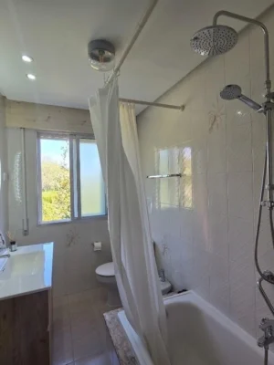 Baño Equipado y Relajante en Casa Buen Camino - Descanso en Arzúa Cuarto de baño moderno en Casa Buen Camino, Arzúa. Dispone de una bañera con mampara blanca, columna de ducha de acero inoxidable con doble salida (cabezal de lluvia y de mano), un mueble de lavabo de madera con espejo circular retroiluminado por LED y una ventana que aporta abundante luz natural.