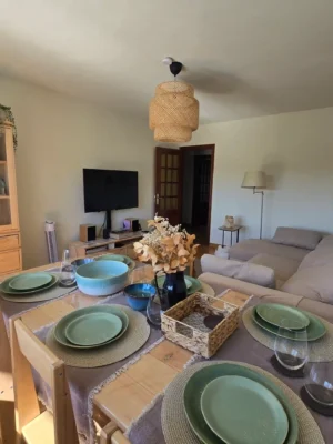 Comedor Equipado con Vajilla en Casa Buen Camino - Tu Descanso en Arzúa Zona de comedor en Casa Buen Camino, Arzúa. En primer plano, una mesa de madera vestida con mantelería gris y juegos de vajilla verdes y azules listos para los huéspedes. A la izquierda, un gran aparador de madera clara con vitrina superior mostrando copas y platos. Al fondo, se aprecia el mueble de televisión y la entrada al salón, con luz natural entrando por la ventana, ofreciendo un ambiente acogedor para dormir en Arzúa y cenar en grupo tras la etapa.