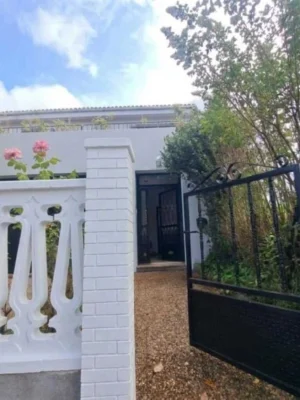 Bienvenida a Casa Buen Camino - Tu refugio en Arzúa Acceso principal a Casa Buen Camino en Arzúa. Se observa una cancela de forja negra abierta, un sendero de grava y una zona ajardinada con flores que precede a la vivienda. El muro blanco con balustrada tradicional refleja la luz natural, ofreciendo una bienvenida serena a los peregrinos del Camino de Santiago.