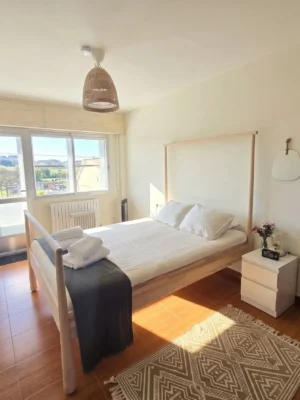 Habitación Doble con Vistas en Casa Buen Camino - Tu Descanso en Arzúa Habitación doble luminosa en Casa Buen Camino, Arzúa. Dispone de una cama con estructura de madera clara, sábanas blancas, toallas limpias y una manta gris. La estancia cuenta con suelo de madera, una gran ventana con balcón con vistas al paisaje verde de Galicia, una mesita de noche blanca con flores, dos espejos decorativos en la pared y una lámpara de techo de mimbre, ideal para el descanso del peregrino en el Camino de Santiago.