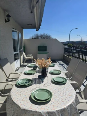 Comedor Exterior Ovalado con Vajilla Completa en Casa Buen Camino - Tu Relax en Arzúa Primer plano de una gran mesa ovalada en la terraza de Casa Buen Camino, Arzúa. La mesa está vestida con un mantel de flores y dispuesta con un juego completo de vajilla en tonos verdes y azules, con copas de vino y un jarrón de flores secas en el centro. Alrededor, sillas de terraza modernas bajo un cielo azul despejado. Al fondo, se aprecia la barbacoa verde y el resto de la terraza, ofreciendo un ambiente exclusivo para dormir en Arzúa y disfrutar de cenas al aire libre tras la etapa del Camino de Santiago.