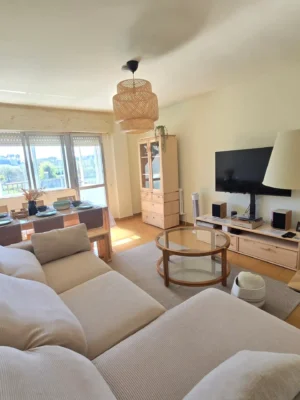 Zonas Comunes: Un espacio diseñado para compartir y relajar Amplio salón comedor de Casa Buen Camino en Arzúa. La estancia cuenta con un sofá tipo chaise longue en tonos beige, mesa de comedor montada para huéspedes, televisión, mobiliario de madera clara y una gran ventana con vistas al paisaje gallego. Decoración acogedora y moderna para el descanso del peregrino.