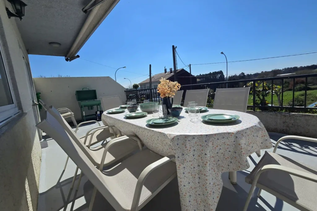 Spacious outdoor terrace at Casa Buen Camino, Arzúa, with a large dining table for groups, comfortable chairs and a green barbecue in the background. The table is set with a floral tablecloth and full tableware, under a clear sky with open views of the Galician countryside, ideal for pilgrims' outdoor dinners.