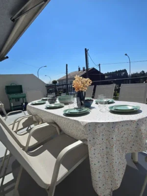 Terraza con Barbacoa y Vistas en Casa Buen Camino - Relax en Arzúa Amplia terraza exterior en Casa Buen Camino, Arzúa, equipada con una mesa grande de comedor para grupos, sillas confortables y una barbacoa verde al fondo. La mesa está vestida con un mantel de flores y vajilla completa, bajo un cielo despejado con vistas despejadas al entorno rural de Galicia, ideal para cenas al aire libre de peregrinos.